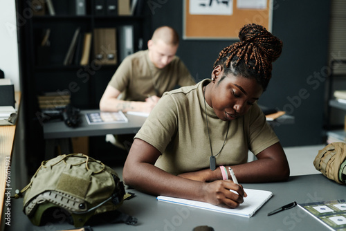 Young African American female student making notes in notepad after teacher while sitting by desk at lesson of military training photo