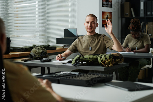 Young male student raising hand and looking at teacher at lesson while asking him question after lecture about military training photo