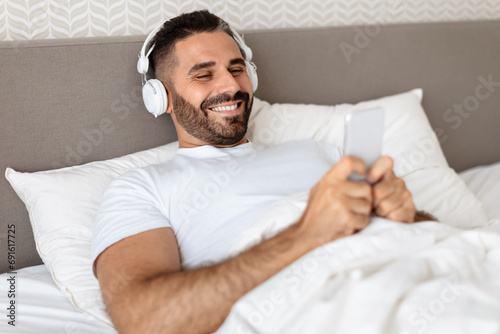 man lying in bed with earphones and phone in bedroom