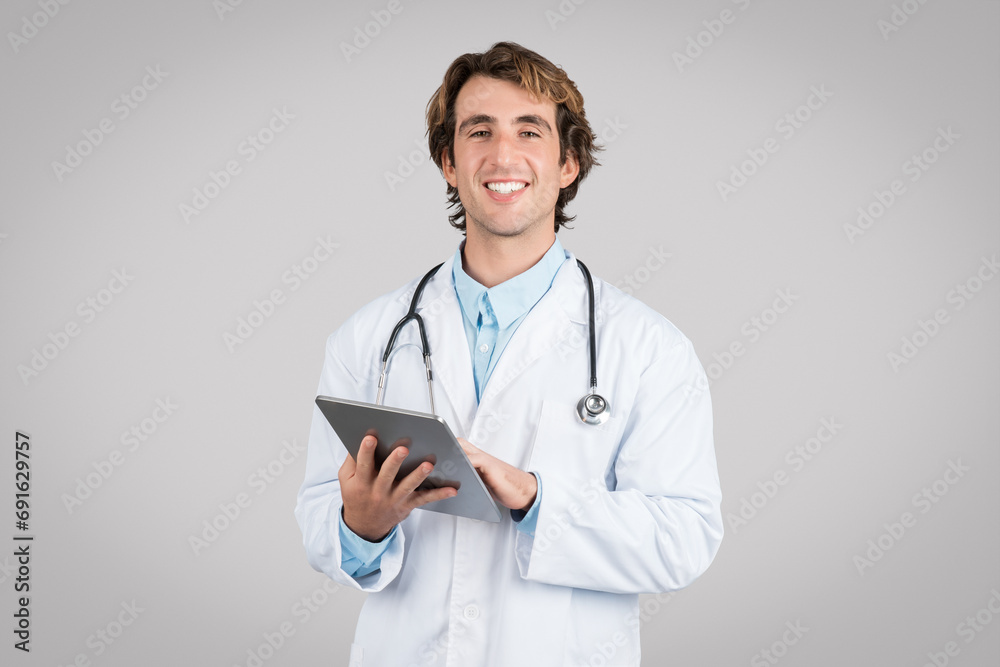 Portrait of cheerful male doctor in white uniform using digital tablet and smiling at camera, consulting patients online Portrait of cheerful male doctor in white uniform using digital tablet and smiling at camera, consulting patients online