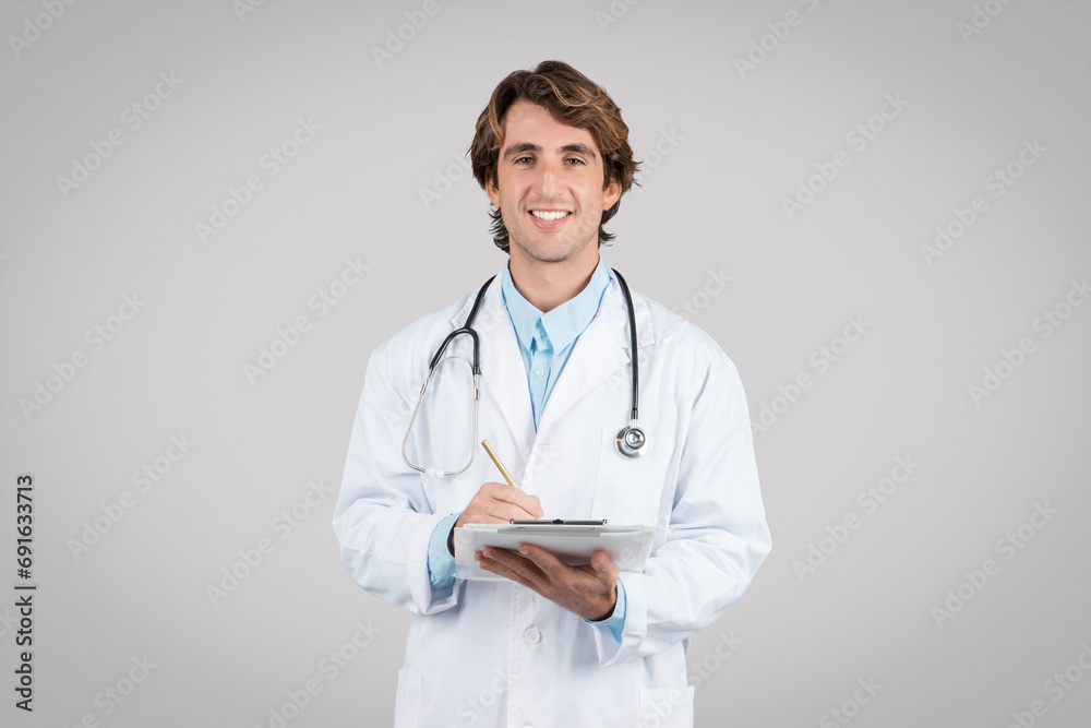 Positive european man doctor therapist in white coat chatting on tablet, smiling at camera isolated on grey background Positive european man doctor therapist in white coat chatting on tablet, smiling at camera isolated on grey background