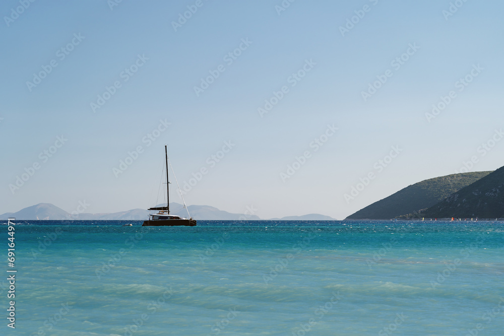 Obraz premium A lone sailboat drifts on the expansive blue waters under the vast sky, framed by distant hills