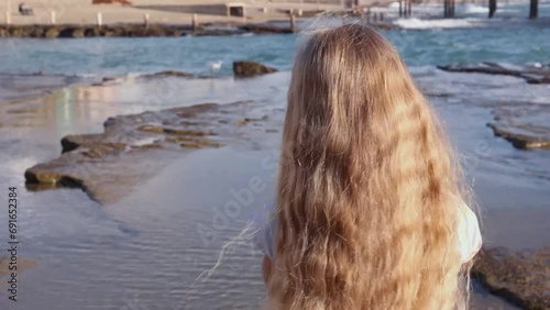 Little girl watches a small white bird walk on sea rocks, beautiful blonde hair blowing in wind