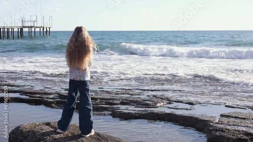 Cinematic seascape, little blonde girl stands on rocks and looks at the wavy sea, girl dreaming and talking to the ocean, wind blows her long hair, feel the sea breeze 