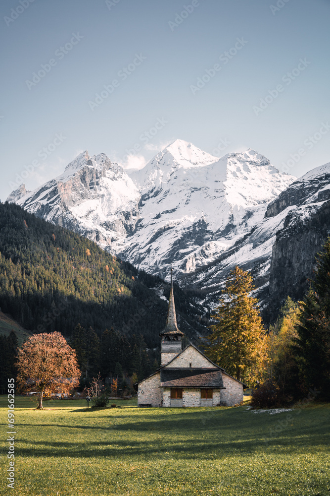 Autumn landscape view of a typical alpine church with colourful trees and snow-capped mountains in the background