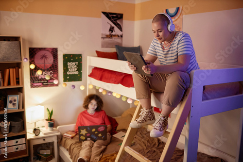Portrait of students dorm room with bunk bed and two young women using tech devices, copy space photo