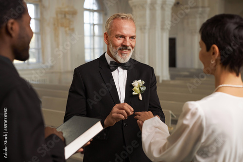 Waist up portrait of smiling senior groom exchanging rings with wife during wedding ceremony at church altar photo
