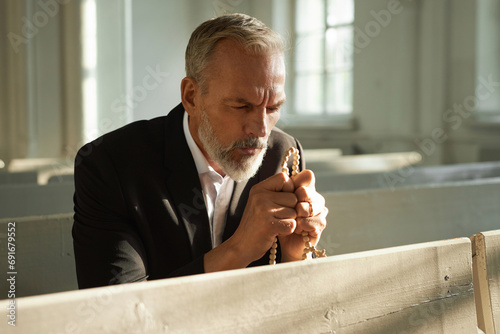 Portrait of bearded senior man praying in church and holding rosary beads, copy space