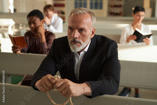 Portrait of bearded senior man praying and holding rosary beads in church on Sunday