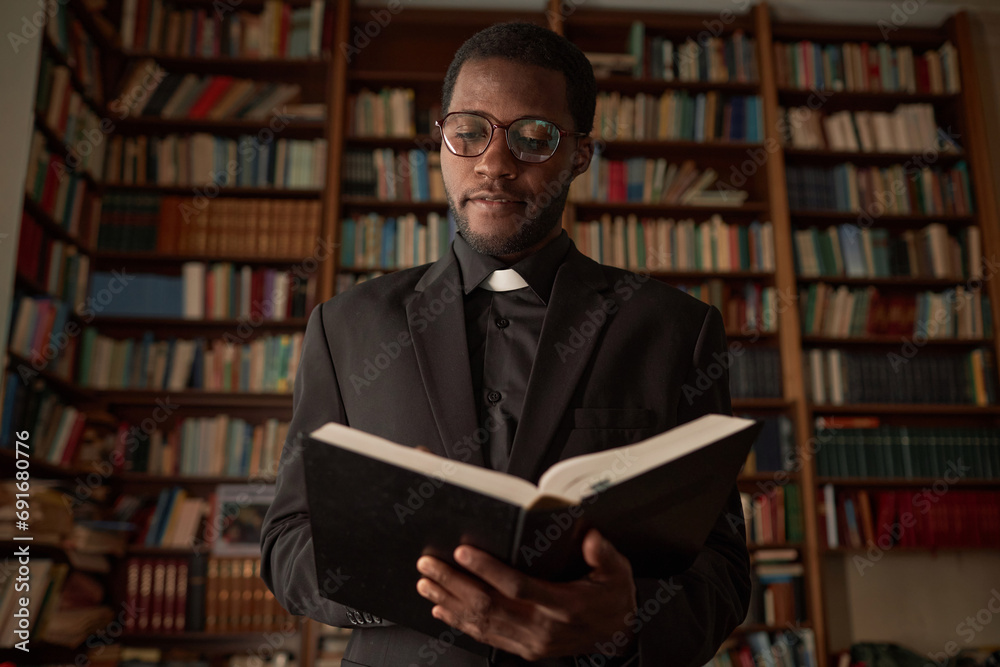 Waist up portrait of young Black man as priest reading Bible in church office with books in background, copy space Waist up portrait of young Black man as priest reading Bible in church office with books in background, copy space