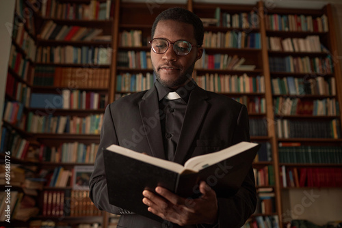 Waist up portrait of young Black man as priest reading Bible in church office with books in background, copy space