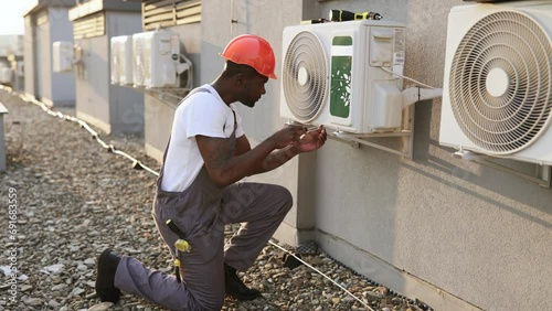 Qualified builder wearing uniform inspecting air conditioner and using screwdriver during repairing work. Black serious male standing on one knee and fixing device in roof of building. photo