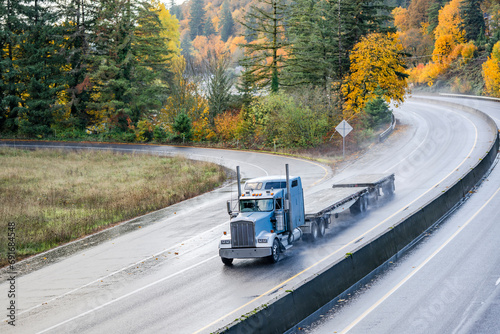 Light blue classic big rig bonnet semi truck tractor transporting two empty flat bed semi trailer driving on the wet autumn highway road with rain dust at rainy weather