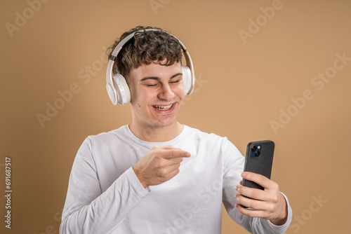 One man caucasian young male stand studio shot on beige background use mobile phone smartphone with headphones listen music send messages texting or browse internet online app copy space © Miljan Živković