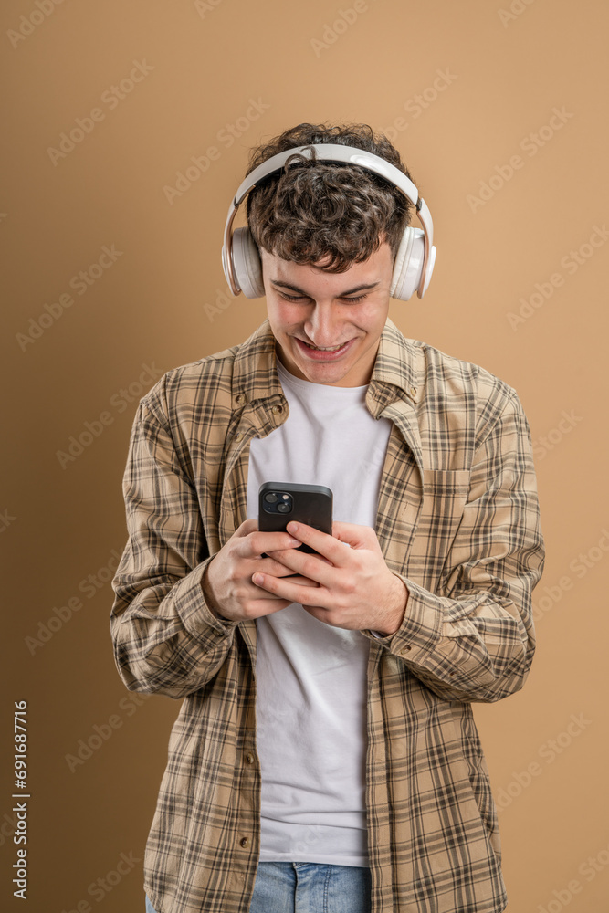 Fototapeta premium One man caucasian young male stand studio shot on beige background use mobile phone smartphone with headphones listen music send messages texting or browse internet online app copy space