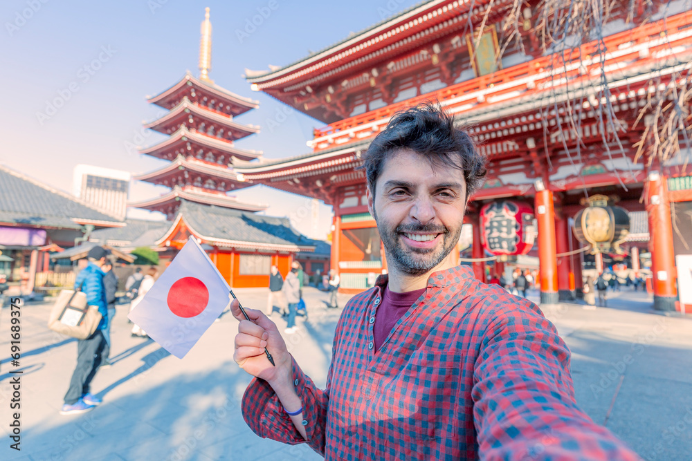 Handsome young tourist enjoying summer holiday in Tokyo, Japan - Traveling life style concept with smiling man taking selfie on city street with japan flag- Tourism and summertime vacation concept - obrazy, fototapety, plakaty 