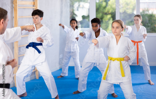 Focused tween girl, karateka beginner in kimono, standing in attacking stance, practicing punches or hand striking techniques as part of kata with group of teenagers.. © JackF