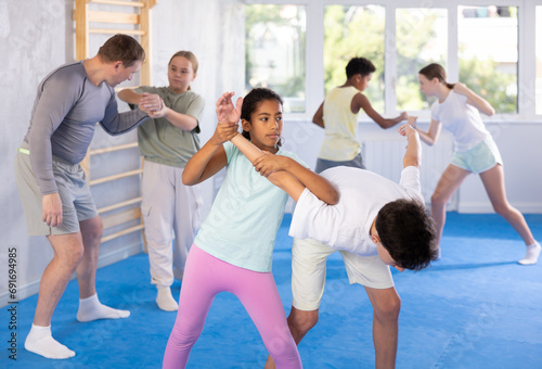 Girls and boys practicing in pair self-defence movements with male trainer supervision photo