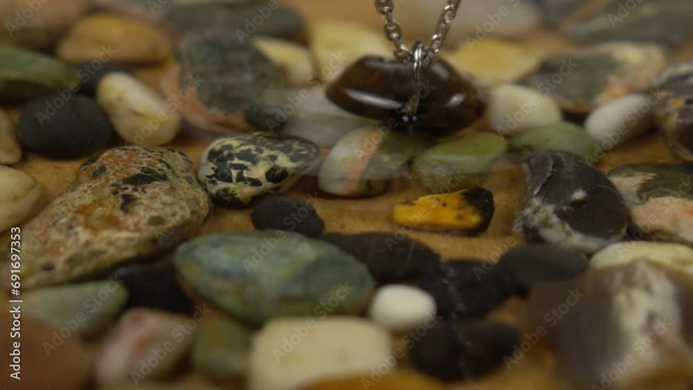 Silver chains with a sea pebble pendant with a hole swings over multicolored pebbles in water, slow motion Silver chains with a sea pebble pendant with a hole swings over multicolored pebbles in water, slow motion