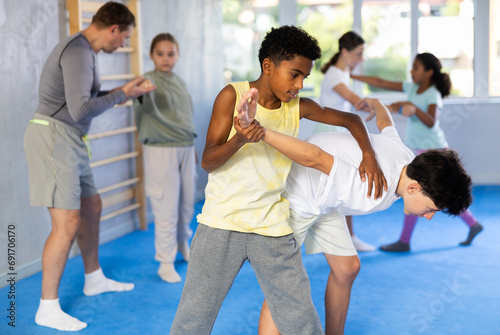 Concentrated african teenager learning effective self defence techniques in sparring with boy in training room, practicing painful armlock .. photo