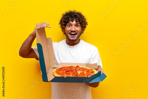 hungry Hindu guy opens a box with delicious pizza on a yellow isolated background, young Hindu man holds fast food photo