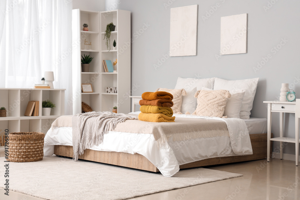 Stack of warm knitted sweaters on bed in modern bedroom - obrazy, fototapety, plakaty 