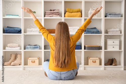 Young woman sitting on floor near shelves with different clothes at home photo