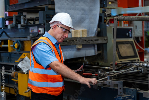 Caucasian industrial worker is using laptop computer to calibrating machine while inspecting inside the metal sheet galvanized roof factory for safety industry