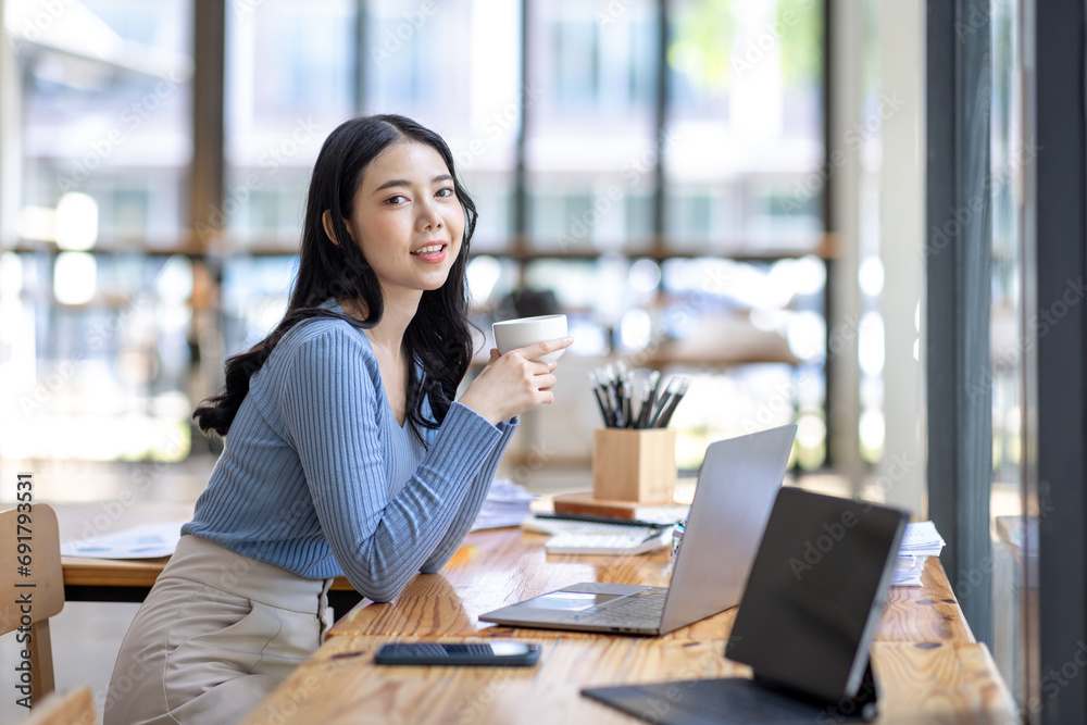 Beautiful young smiling Asian businesswoman working on laptop and drinking coffee, Asia businesswoman working document finance and calculator in her office.
 - obrazy, fototapety, plakaty 