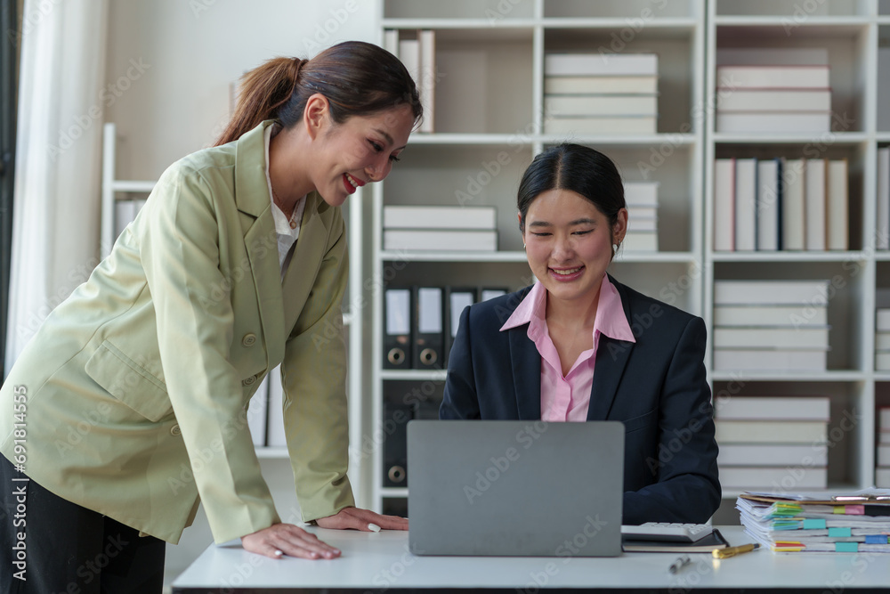 Fototapeta premium Two Asian office women cooperate and encourage each other sitting at desks with laptops. Discuss financial management planning Analysis of startup project ideas and office investment market statistics