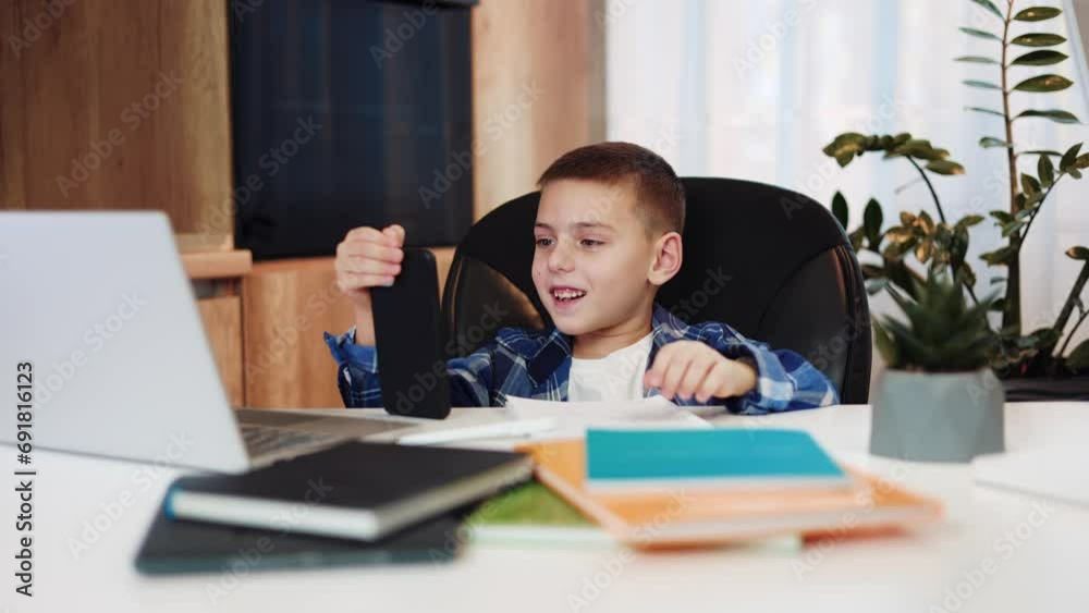 School kid having video call on modern smartphone during distance education in front of desk with computer and plants. Child using technology for doing online school program at cozy apartment. School kid having video call on modern smartphone during distance education in front of desk with computer and plants. Child using technology for doing online school program at cozy apartment.