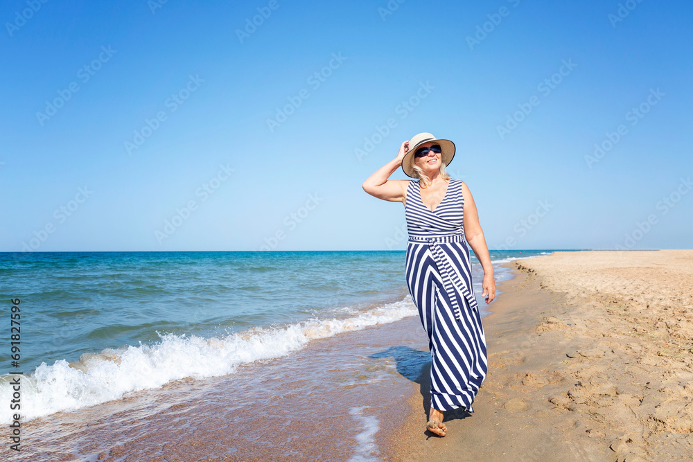 A smiling elderly woman walks along the seashore. Senora in a long striped dress and hat. Active recreation and relaxation at the resort. A smiling elderly woman walks along the seashore. Senora in a long striped dress and hat. Active recreation and relaxation at the resort.