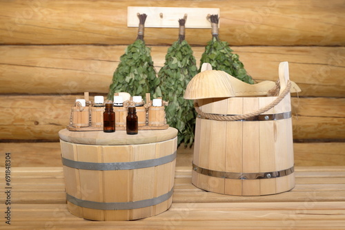 Bottles of healing essential oil are on cover of a wooden vat standing next to wooden bucket with ladle against the background of dry birch whisk brooms hanging on a log wall.