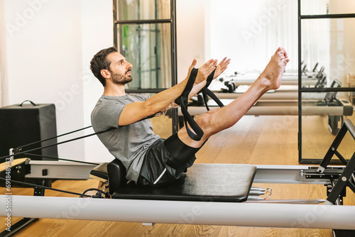 Young man in sportswear doing teaser Pilates exercise and stretching on reformer bed photo