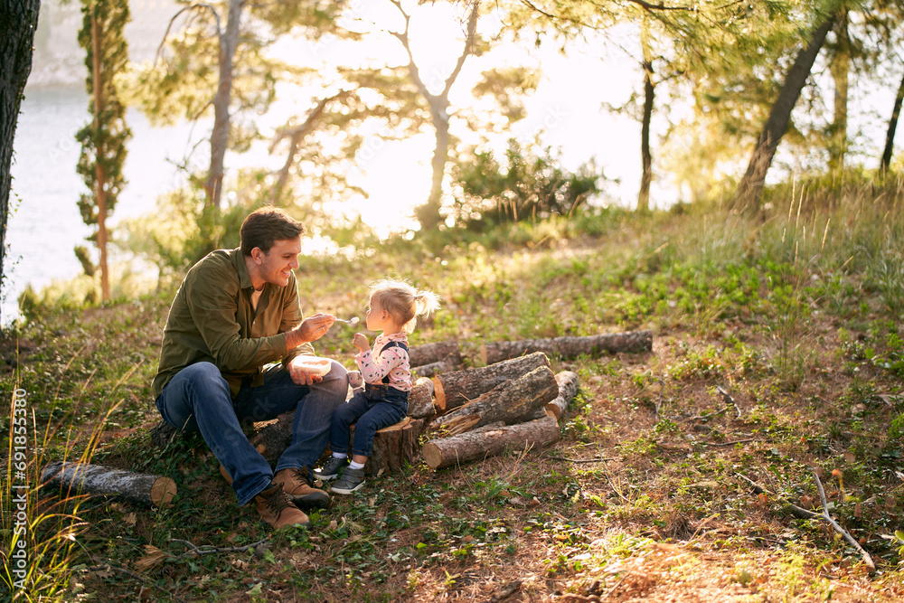 Dad is spoon-feeding porridge to a little girl sitting on logs in a sunny forest