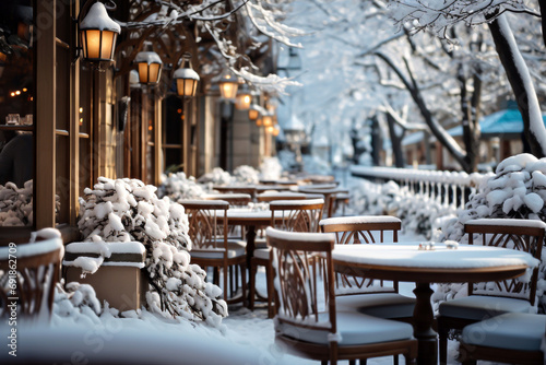 Tables and chairs of outdoor restaurant during winter snowfall. Defocused lanterns lights