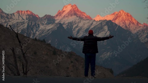 Rear view shot of an Indian man wearing hat and jacket raising arms in front of snow covered Himalayan mountain peaks during the sunset at Kinnaur in Himachal Pradesh, India. Traveler raises his arms photo