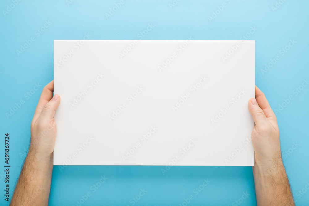 Young adult man hand holding and showing new white ceramic tile on light blue table background. Pastel color. Closeup. Point of view shot. Empty place for text. Top down view. - obrazy, fototapety, plakaty 