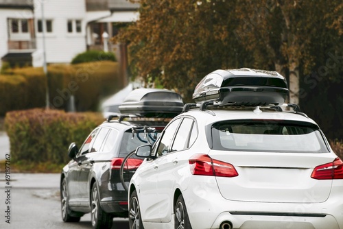 Rooftop cargo carrier bag. Rear view of a car with a roof box. Parked cars in Europe. Black Roof Box on a Sporty White Wagon Family Car. Removable black car trunk for luggage on the roof of a car.