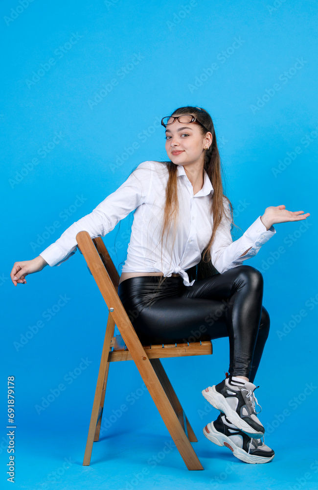Attractive woman with glasses on forehead, smiling at camera, while sitting on chair. Side view of pretty girl in black leggings turning to camera, while posing, isolated on blue. Concept of posing. Attractive woman with glasses on forehead, smiling at camera, while sitting on chair. Side view of pretty girl in black leggings turning to camera, while posing, isolated on blue. Concept of posing.