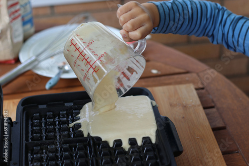Fototapeta Naklejka Na Ścianę i Meble -  Child making waffle with the waffle machine plate. Child holding cup pouring wet cake mixed batter in a waffle machine for his breakfast. Family Cooking concept