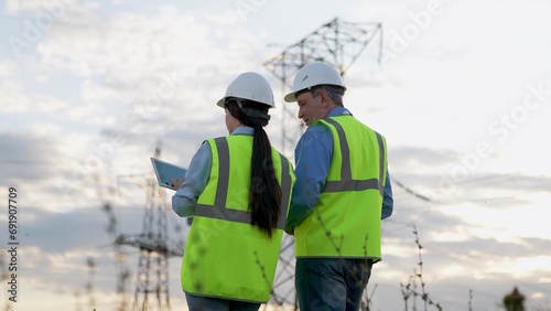 Engineer demonstrates electric power distribution substation to colleague with tablet at sunset. Electricians inspect equipment with tablet at power generation plant with transmission lines in field