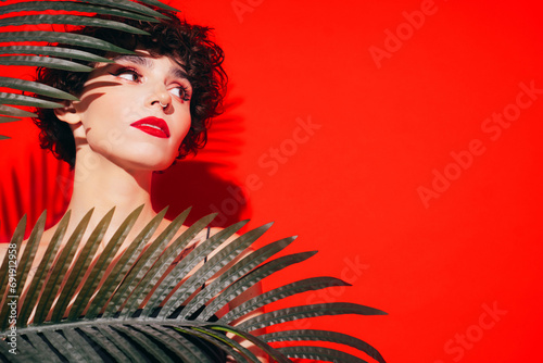 Fashion beauty portrait of young confident brunette woman with evening stylish makeup and perfect clean skin. Sexy model with curly hair posing in studio. With red bright lips. Tropical palm leaf