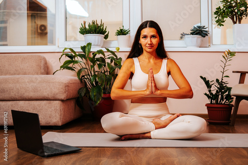 Beautiful young brunette woman is sitting in lotus position with her hands in namaste on yoga mat at home in living room.