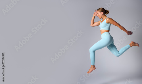 Studio Shot Of Woman Wearing Gym Fitness Clothing In Mid-Air Exercising On Grey Background
