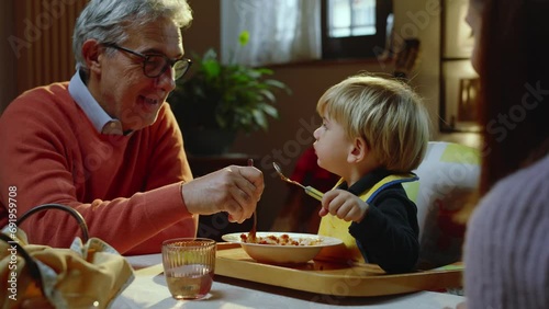 A young grandfather feeds pasta to his beautiful grandson while sitting at the dining table together with the rest of the family