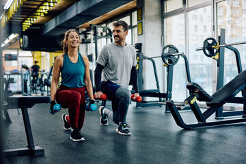 Happy athletic couple exercising with hand weights in lunge position in gym.