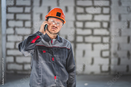 Bearded construction caucasian worker in boilersuit, helmet and protective glusses talking on a smartphone at a construction site. photo