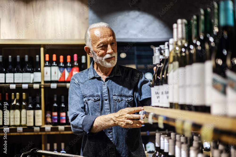 Male consumer buying bottle of red wine in winery store. Smiling mature man wearing casual clothes shopping at supermaket grocery store buy choosing wine alcohol hold bottle inside hypermarket.