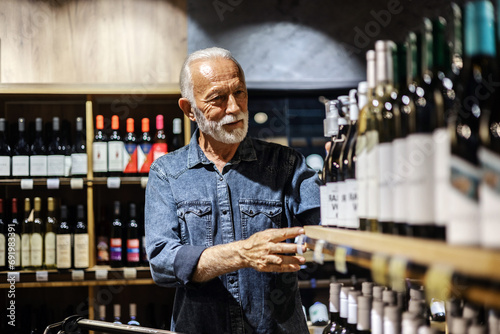 Male consumer buying bottle of red wine in winery store. Smiling mature man wearing casual clothes shopping at supermaket grocery store buy choosing wine alcohol hold bottle inside hypermarket. photo